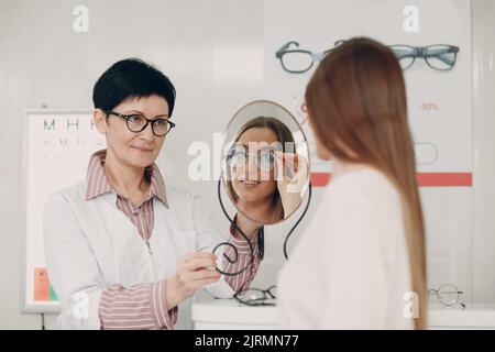 Young smiling woman smiling picking and choosing glasses at the optician corner at the shopping mall at mirror. Happy beautiful woman buying eyewear eyeglasses at the optometrist. Stock Photo