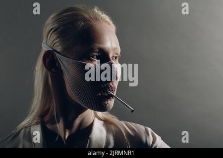 Closeup portrait of white caucasian albino blond woman fashion model wearing quarantine medical face mask coarse mesh net with cigarette on grey background Stock Photo