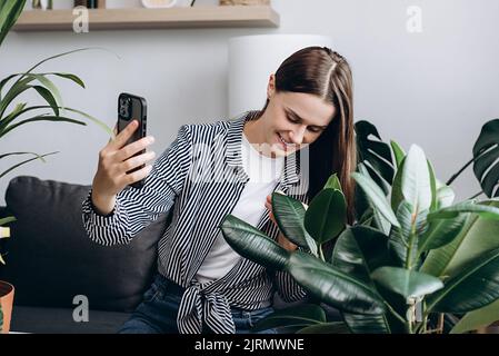 Happy beautiful young female florist blogger being photographed with plants in pot on mobile phone camera. Home plant breeding, gardening, housewife, Stock Photo