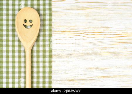 Wooden spoon with smile on green checkered tablecloth on white wooden surface. Mockup for menu or recipe, restaurant, website with cooking. Kitchen fo Stock Photo