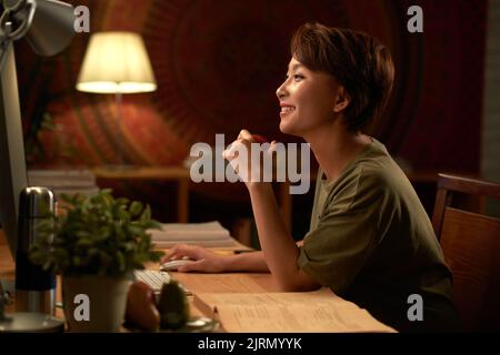 Side view of cheerful young Asina woman reading something funny on computer screen Stock Photo