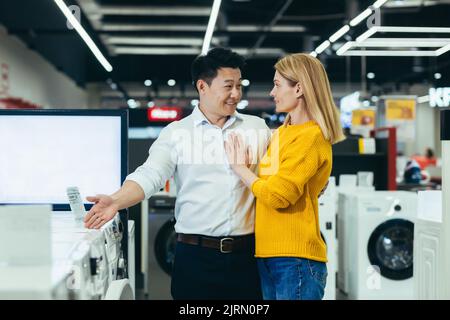 Happy diverse young family man and woman choose electronic household appliances, shop for new apartment in supermarket Stock Photo