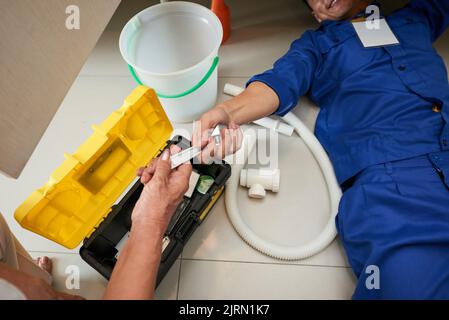 Senior woman passing wrench to plumber who is lying on the floor Stock Photo