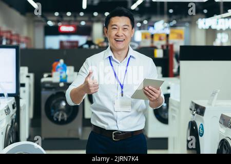 Successful sales consultant in electronics supermarket, Asian man working at home appliances store smiling and looking at camera, man holding tablet portrait and showing thumb up recommending Stock Photo