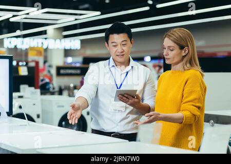 Asian consultant salesman in electronics and household appliances store, selling a working machine to a woman, recommending and approving the choice Stock Photo