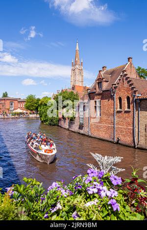 The spire of the Church of Our Lady (Onze-Lieve-Vrouwekerk) overlooking tourists enjoying a boat trip and old houses beside the canal in Bruges, Belgi Stock Photo