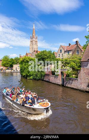 The spire of the Church of Our Lady (Onze-Lieve-Vrouwekerk) overlooking tourists enjoying a boat trip and old houses beside the canal in Bruges, Belgi Stock Photo