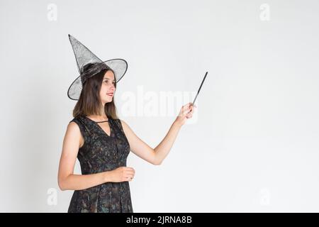 A beautiful cute girl in a witch costume, wearing a hat, with a magic wand shows an empty space on a white background, copy space. The sorceress. Happ Stock Photo