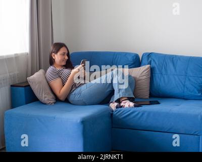 Woman lying on blue sofa in her living room and reading the news on a smartphone Stock Photo