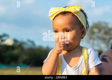 close-up of a beautiful brown-skinned latina baby girl with one hand on her mouth smiling brightly in an outdoor field on a beautiful summer day. Stock Photo