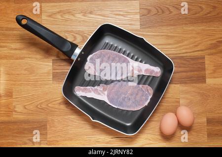 Rashers of raw bacon placed in a griddle frying pan, two brown hens eggs placed at the side on a wooden table top. Shot from above Stock Photo