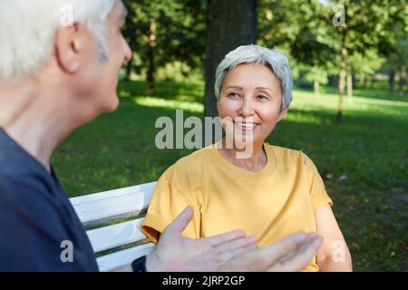 Happy older couple talk sit on bench in summer park, wear sportive clothes, enjoy conversation in the morning outdoors, focus on wife listen husband. Stock Photo