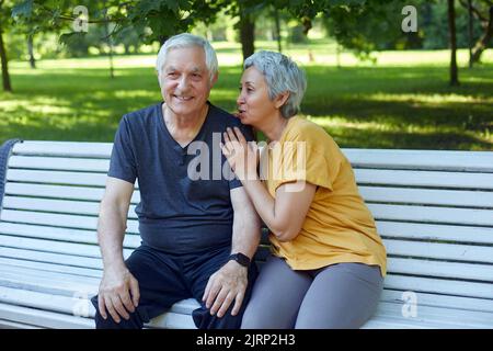 Attractive older couple talking seated on bench in summer park, wear sportive clothes, enjoy conversation in the morning outdoors. Communication, harm Stock Photo