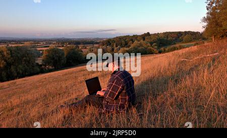 Man using laptop sitting on a hill in the countryside Stock Photo