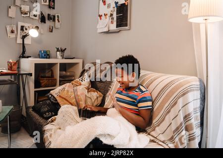 Black boy gesturing and using laptop while sitting on sofa at home Stock Photo
