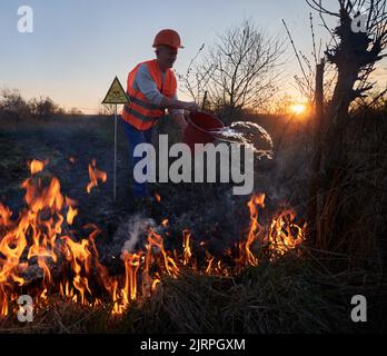 Fireman ecologist extinguishing fire in field in the evening. Male environmentalist holding bucket and pouring water on burning dry grass near yellow triangle with skull and crossbones warning sign. Stock Photo