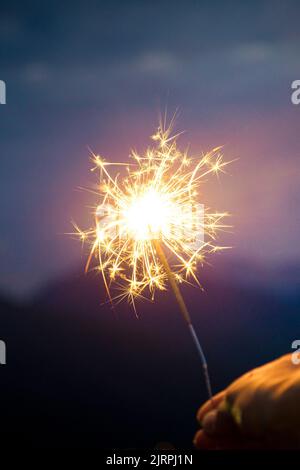 Hand holding a sparkler, or bengal fire stick, burning in outdoor setting, mountain landscape, at dusk. Holidays or magic background or wallpaper. Ver Stock Photo