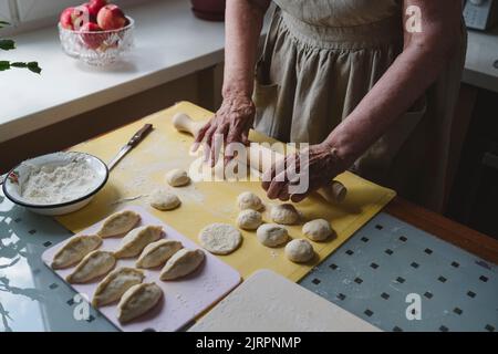 An elderly woman rolls out the dough with a rolling pin on pies. Stock Photo