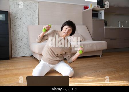 Active caucasian senior woman doing fitness at home on the floor in front of a laptop. Mature lady takes care of her healthy body and mind by Stock Photo