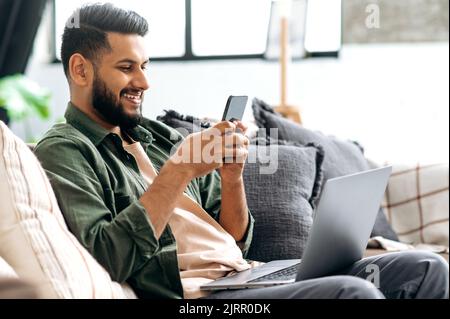 Positive handsome indian or arabian guy, sit in a modern living room on comfortable sofa, uses smartphone, texting message, browsing social media, surfing web, smiles. The concept of using gadgets Stock Photo
