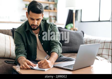 Concentrated clever indian or arabian guy, student or freelancer, sitting at home on sofa in living room, using laptop, working or studying remotely, listening to webinar, taking notes, homework Stock Photo