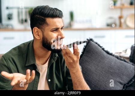 Speakerphone conversation. Positive indian or arabian young man in casual wear, sit on a sofa in a cozy living room, holding his smartphone, talking on speakerphone and smiling, record voice message Stock Photo