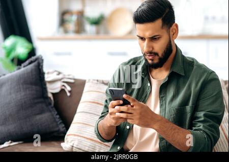 Puzzled indian or arabian guy in casual wear, sits on sofa in a living room, using smartphone, texting online, browsing social networks, reads news, looks at screen in confusion. Wireless technologies Stock Photo