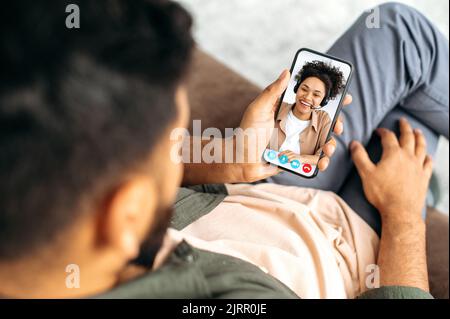 Online video consultation. On a screen of the phone, an African American lovely positive girl holds consultation. Husband and wife or friends talk on online video conference using mobile phone, smile Stock Photo