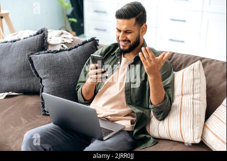 Positive mixed race young man in casual clothes, spending time at home, sits on the sofa in the living room, using a smartphone, read unexpected news, browsing the internet, social networks, smile Stock Photo
