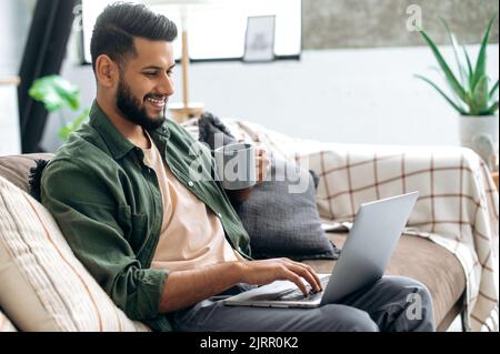 Happy attractive young indian or arabian stylish man, sits on a sofa at home in the living room, using a laptop, browsing the Internet, working on a project with cup of coffee, looks at screen, smile Stock Photo