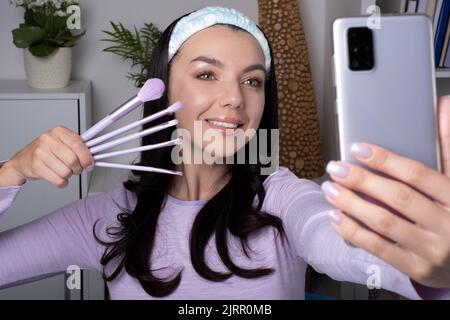 influencer, beauty blogger making a selfie on a mobile phone front camera. an attractive Young woman holds a various cosmetic brush for online review. Stock Photo