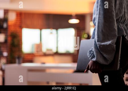 African american female student carrying laptop in home room with modern interior. Young businesswoman in casual clothes holding portable computer, side view with blurred background and copy space Stock Photo