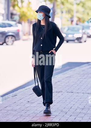 City woman, on a sidewalk, with a mask on her face and walking down the road. Urban fashion, casual walk and covid protection, life in the new normal Stock Photo