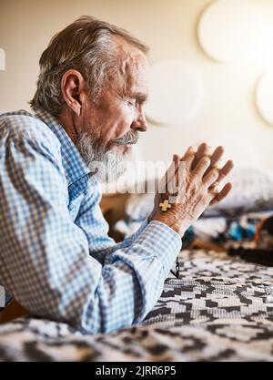 I have so much to be thankful for. a peaceful senior man kneeling next to his bed and praying with his eyes closed. Stock Photo