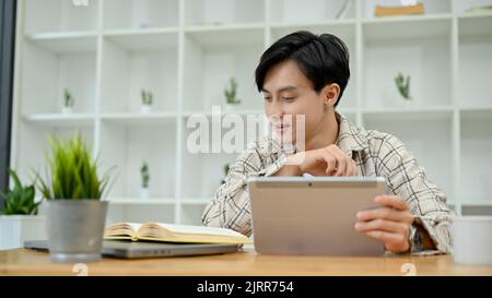 Young Asian male freelancer or college student using digital tablet touchpad while reading some information on the book. Stock Photo