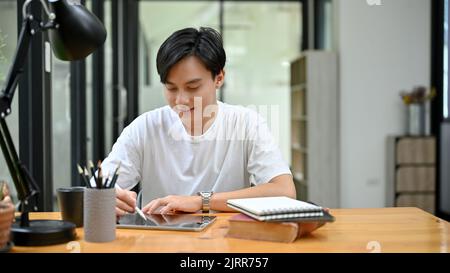 Handsome young Asian male university student concentrating doing his homework on tablet touchpad in the library co-working space. Stock Photo