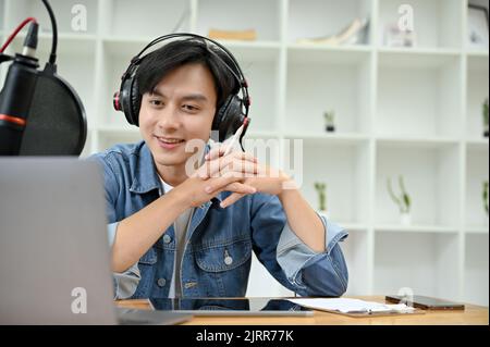 Handsome young Asian man radio host recording and broadcasting his podcast from studio. A man in headphones using microphone and running his radio sho Stock Photo