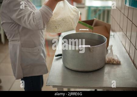 Cook prepares meals. Large pot on table. Cottage cheese in bag. Work as cook in dining room. White coat. Stock Photo