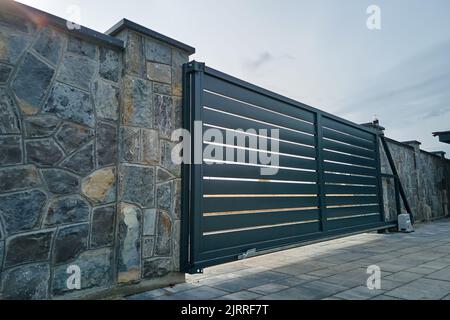 Wide automatic sliding gate with remote control installed in high stone fense wall. Security and protection concept Stock Photo