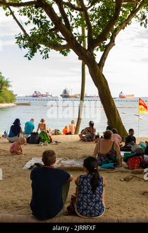Relaxing on Siloso Beach with container ships anchored offshore, Sentosa Island, Singapore Stock Photo
