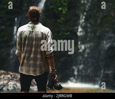 This is amazing. a young man staring at a waterfall with his back turned and holding his camera. Stock Photo