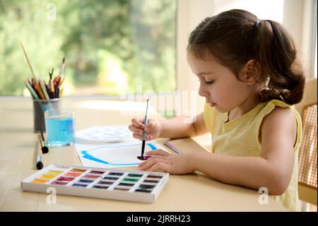 Little girl drawing picture while learning watercolor painture. Colorful palette and paintbrushes on the table Stock Photo