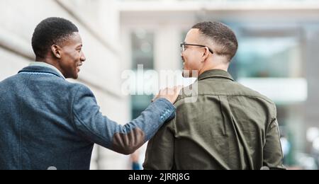 Two young businessmen smiling, talking and walking through the city together, excited for their new project. Happy multiracial coworkers strolling Stock Photo