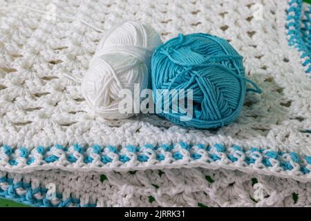 Front view of two skeins, one white and one blue, metallic pink crochet hook on white crochet blanket, blue details on edges, blurred background, rais Stock Photo