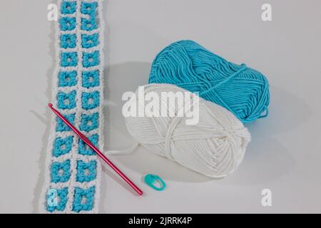 Crochet fabric interlacing the colors white and blue next to two cotton balls, a pink crochet hook and green stitch markers against a white background Stock Photo