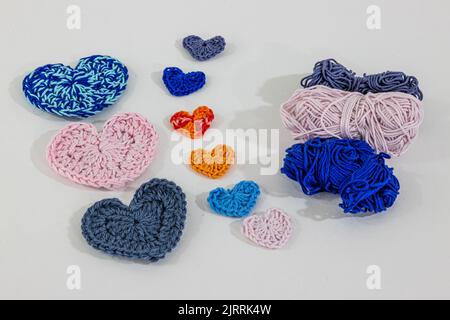 Set of handmade crochet hearts of different colors and sizes next to blue, pink and gray cotton balls on white surface and background, in orange and m Stock Photo