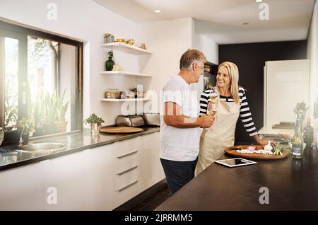 Mature couple cooking, drinking wine and preparing a meal together while bonding, having fun and talking in home kitchen. Smiling, happy and caring Stock Photo
