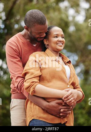 Romantic, loving and caring husband hugging his wife from the back in nature while on a date. Young, African and happy couple embracing each other Stock Photo