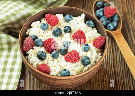 cottage cheese with raspberries and blueberries in brown clay bowl and wooden spoon on brown wooden background. Dairy products, healthy food. Stock Photo