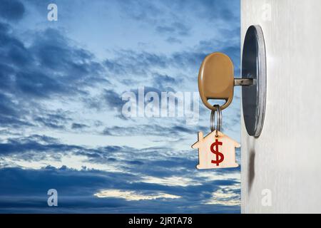 Key with a trinket in the form of a house in the door lock of an open door against the background of the sky with clouds Stock Photo